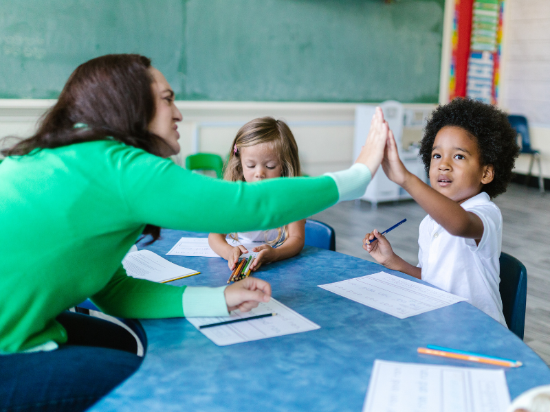 Young student high-fiving a teacher during a classroom activity while another child completes a worksheet.