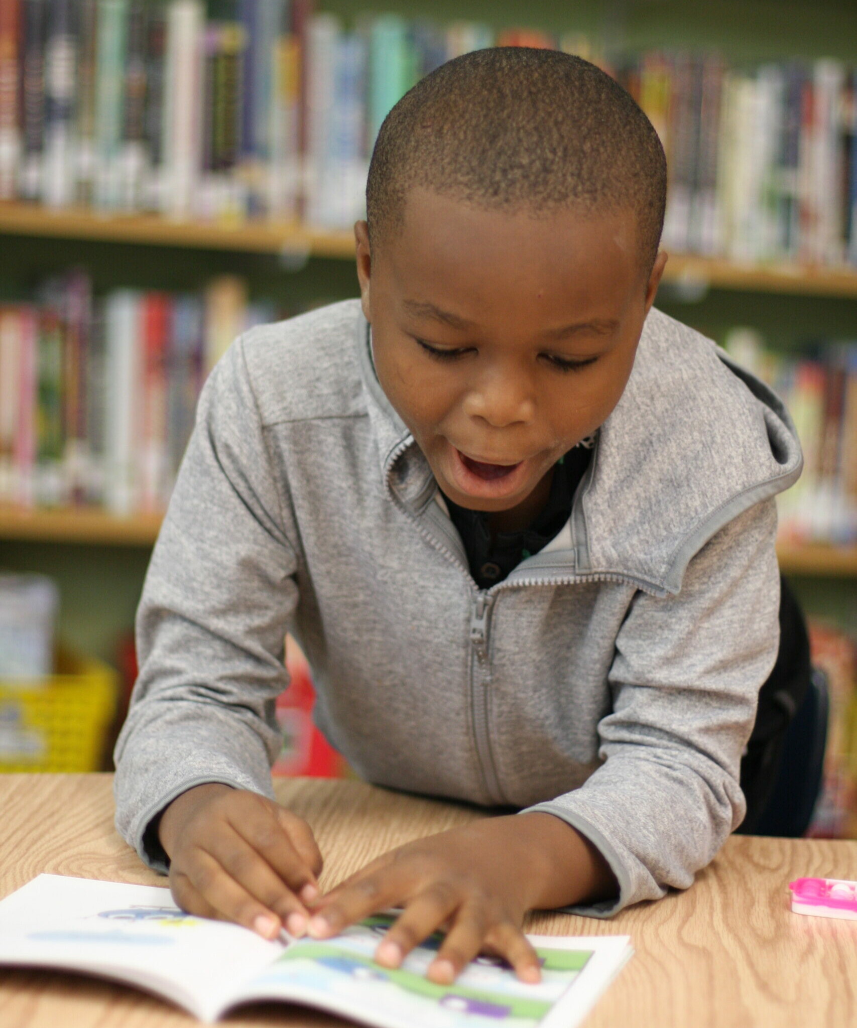Young student reading a picture book at a table in a school library with bookshelves in the background.