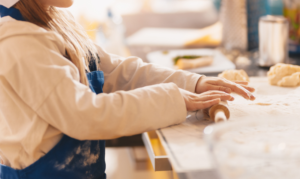 Child wearing an apron rolling dough with a small rolling pin on a floured kitchen counter.