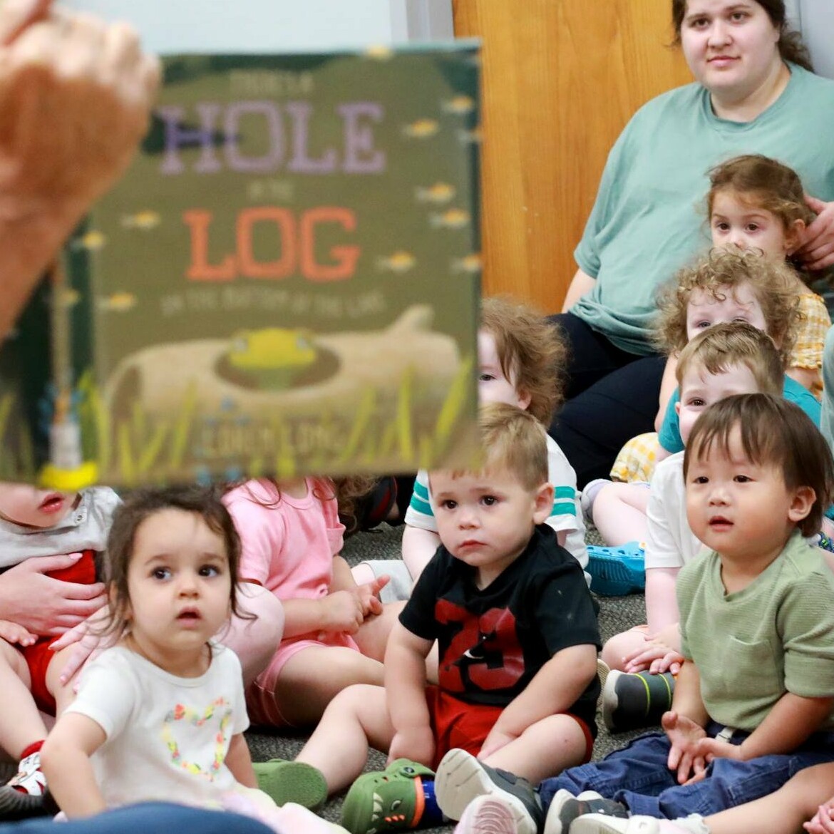 Children sitting on the floor listen attentively as a book is being read to them.