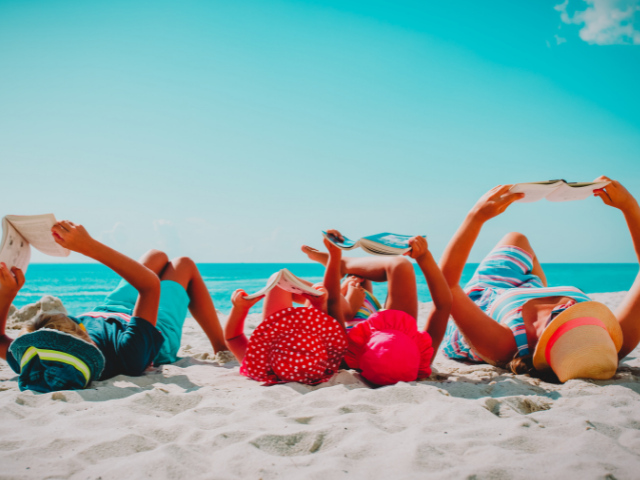 Children lying in the sand at the beach holding books above them on a sunny day by the ocean