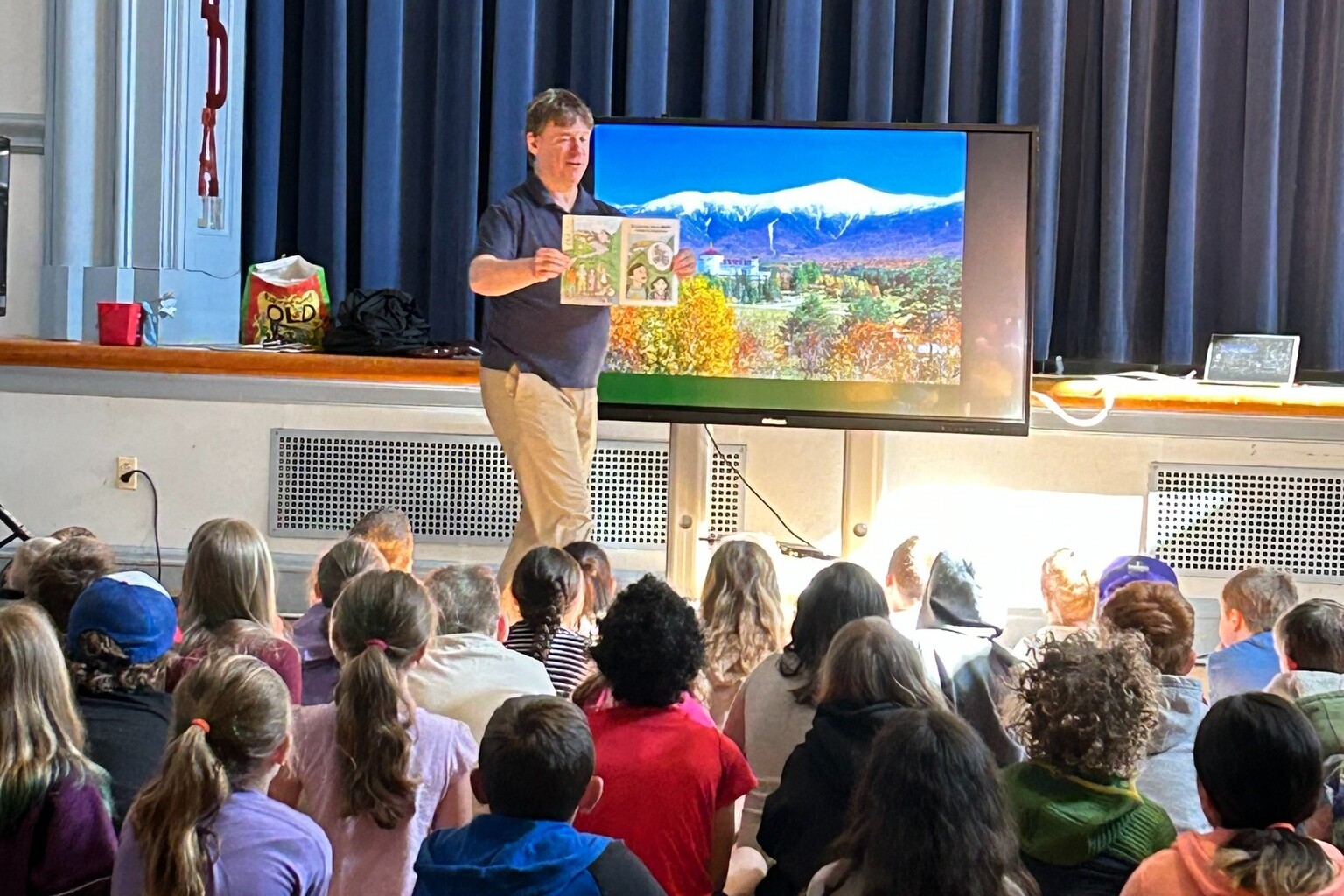 A speaker shows a book to an audience of attentive children in a school setting.