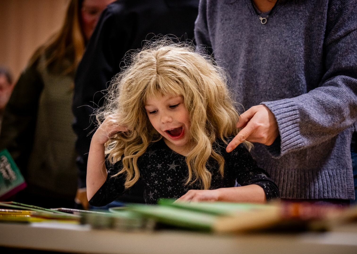 A child with curly hair excitedly examines books on a table.