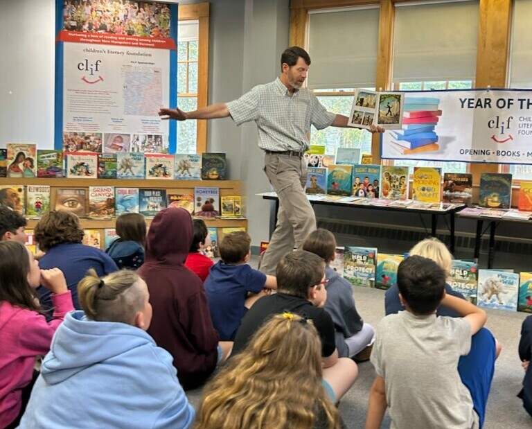 Children sitting on the floor listening to a storyteller presenting illustrated books at a classroom literacy program.