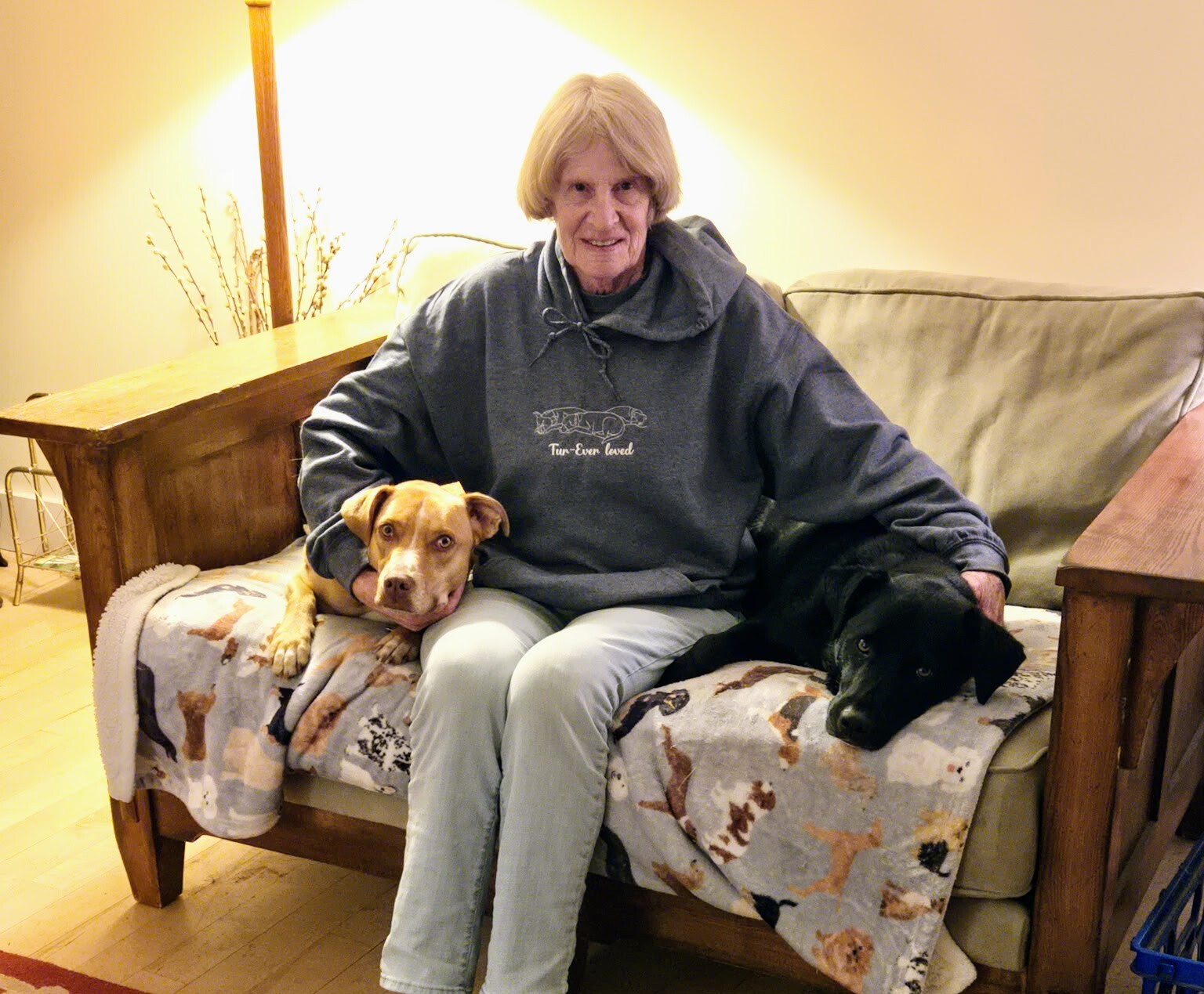 A woman with short hair sits on a couch with two dogs, a tan and a black one.