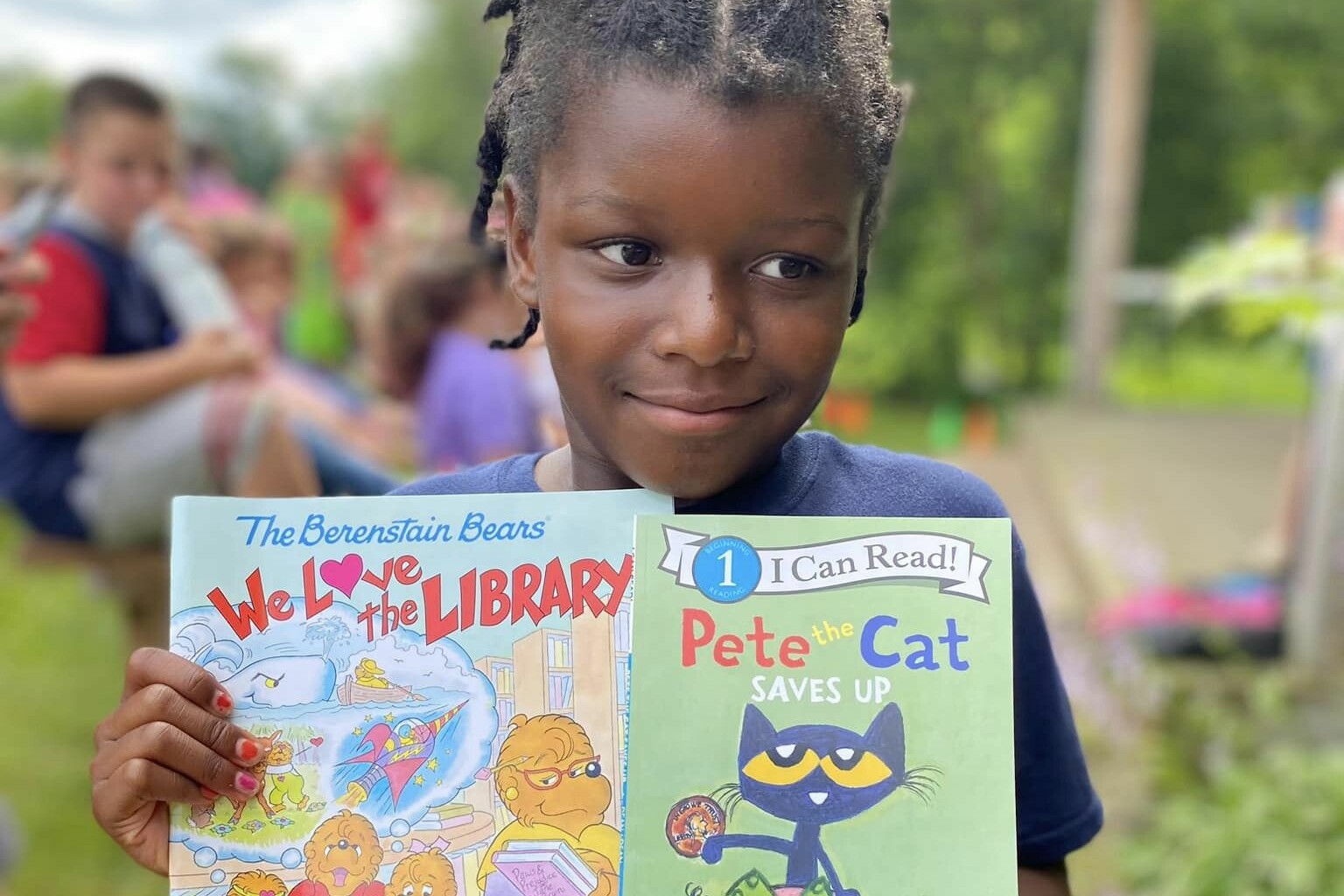 A child holds two books: "We Love the Library" and "Pete the Cat Saves Up" outdoors.