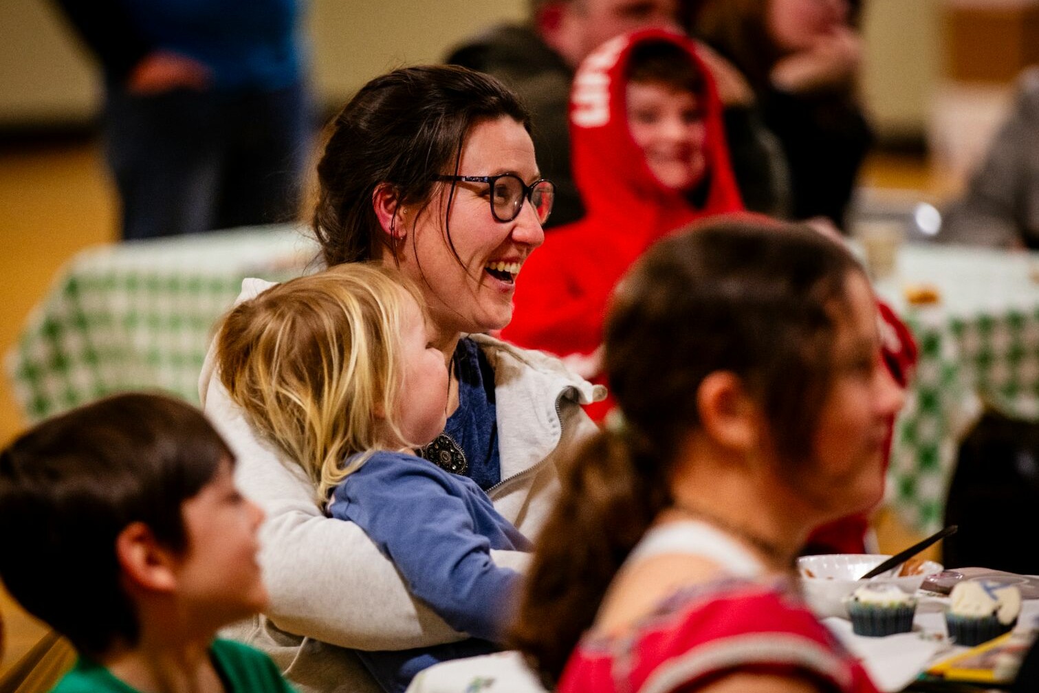 A woman laughs while holding a child, surrounded by smiling people at a gathering.