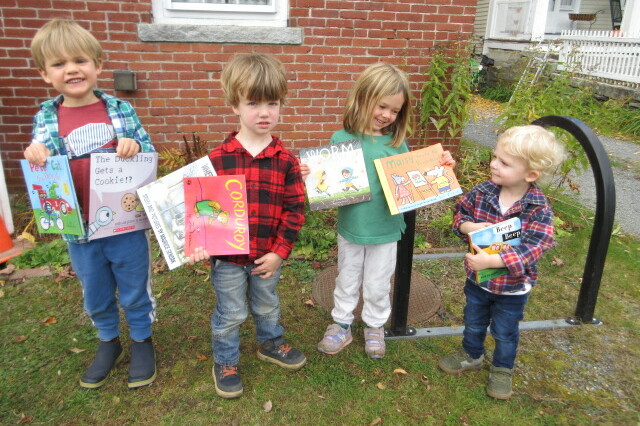 Four children hold up books outside a building, smiling and dressed in colorful outfits.