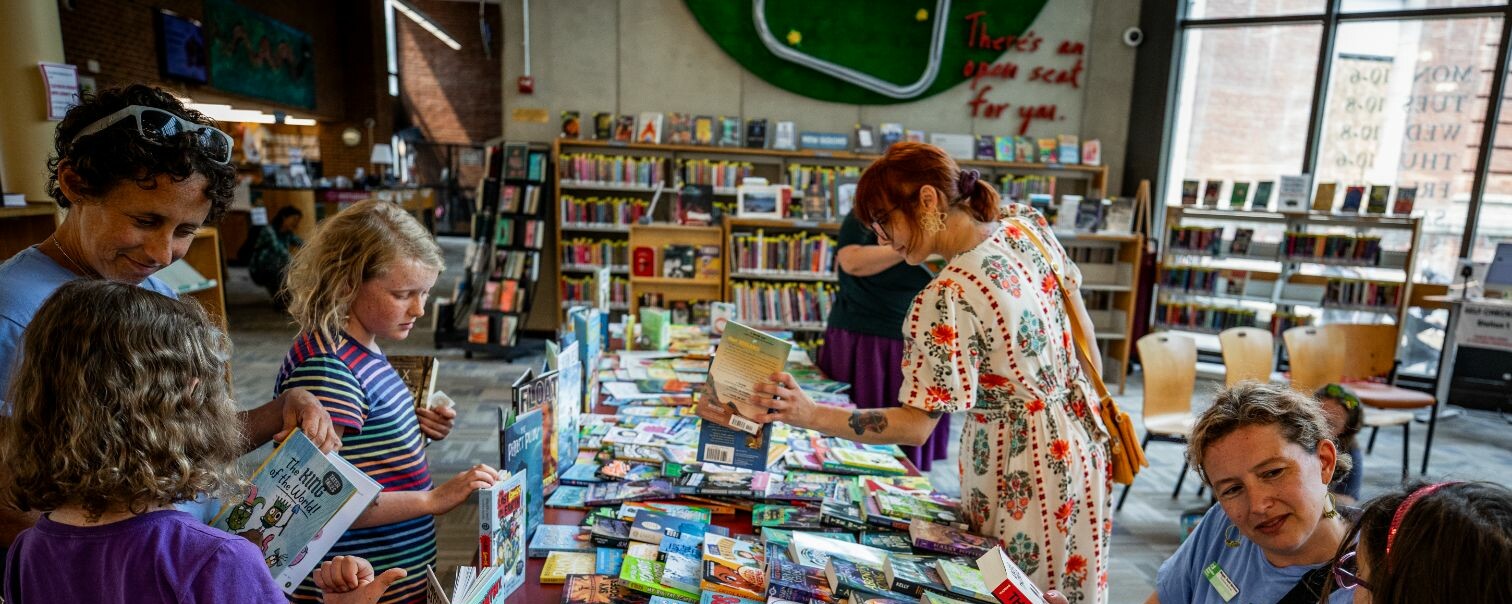 People browsing books at a library table filled with colorful titles.