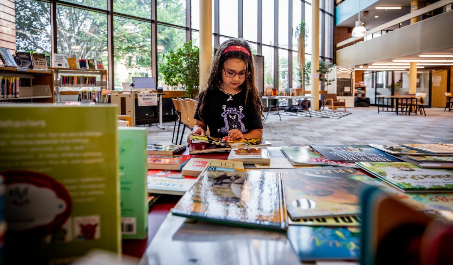A child reads books at a library table surrounded by shelves and natural light.