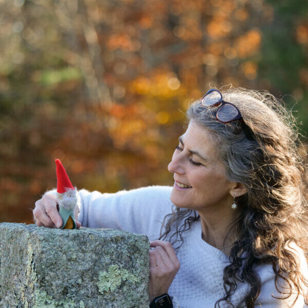 Woman smiles at a small gnome figurine on a stone, with autumn foliage in the background.