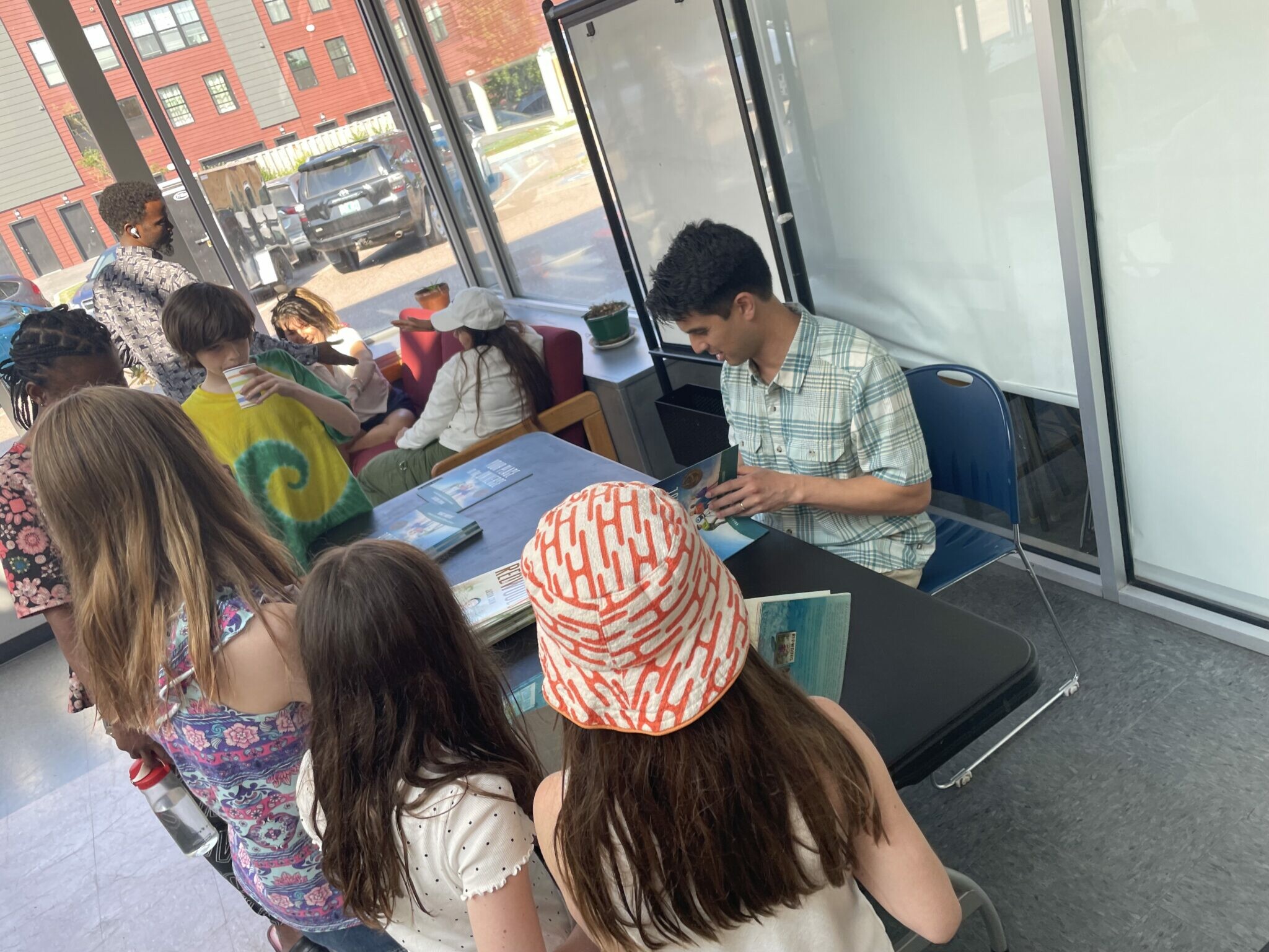 A group of children gathers around a table while a youth looks at a booklet.