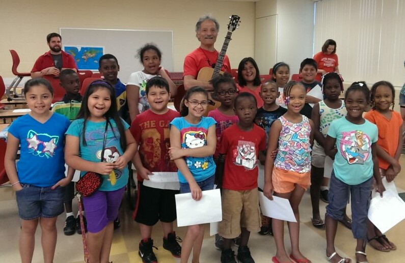 Group of children with a guitar player in a classroom, smiling and holding papers.