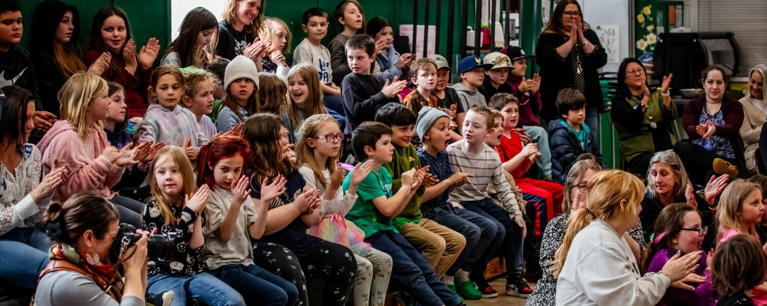 A diverse group of children and adults clapping and enjoying a performance in a school setting.