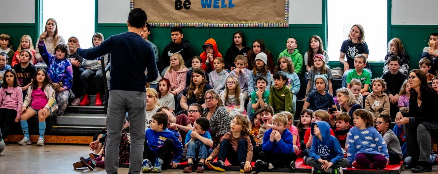A crowd of children attentively listening to a speaker in a gymnasium setting.