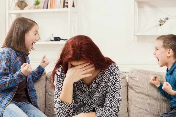 Overwhelmed mother with hands on face as two kids shout beside her on a living room couch
