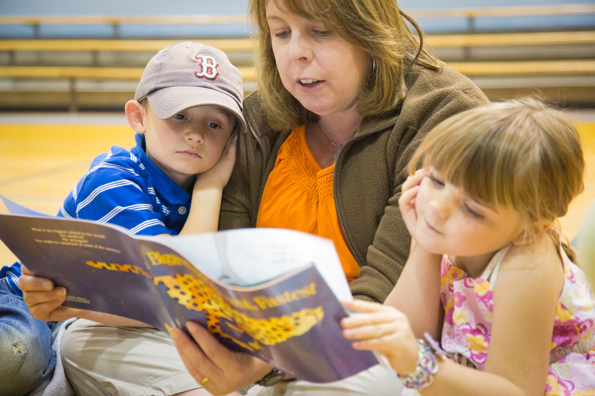 A woman reads a book to two attentive children in a gym setting.