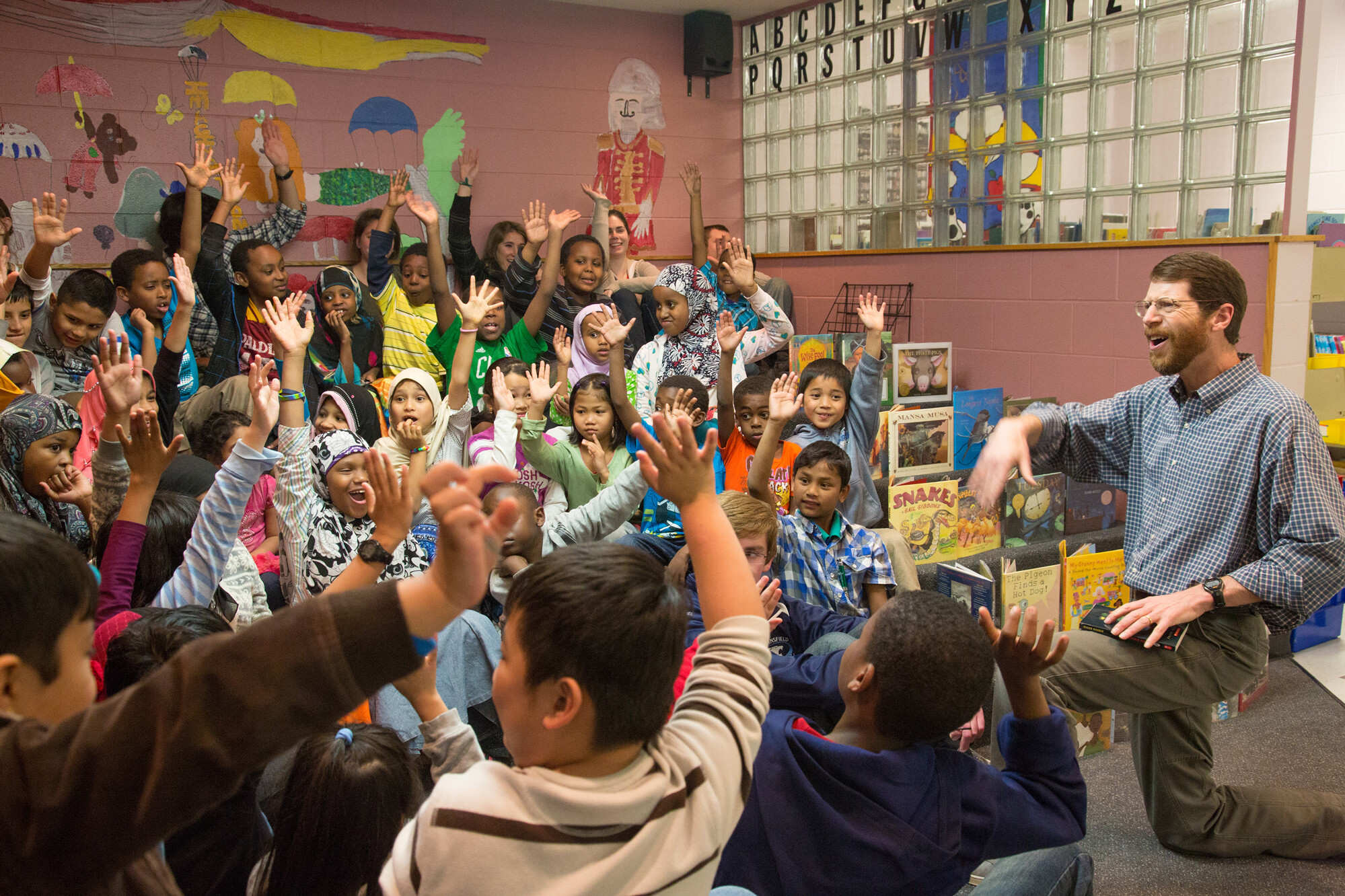 A lively group of children enthusiastically raises their hands during a storytelling session.