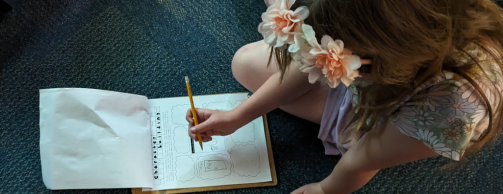 Young girl drawing in a notebook with a pencil while wearing flower hair clips, sitting on a carpeted floor.