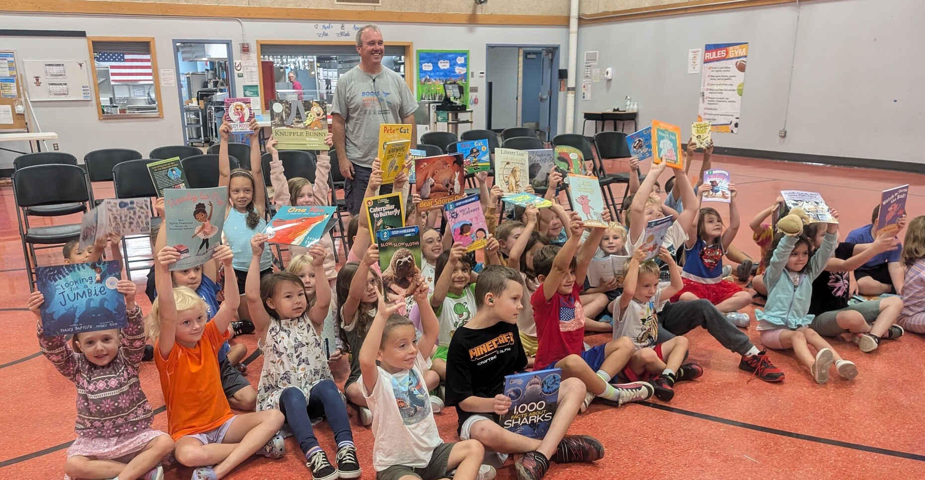 Children hold up books while seated on the floor, smiling with a teacher standing behind them.
