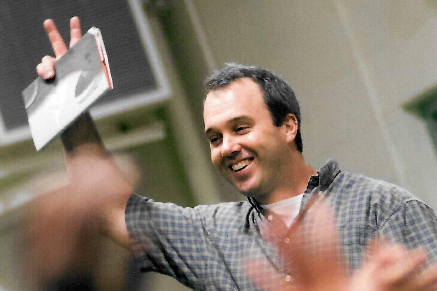 Smiling man holds a book while making a peace sign in a lively setting.