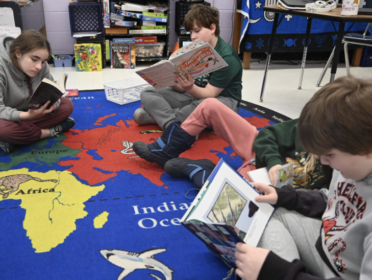 Four children read books while sitting on a colorful map carpet in a classroom.