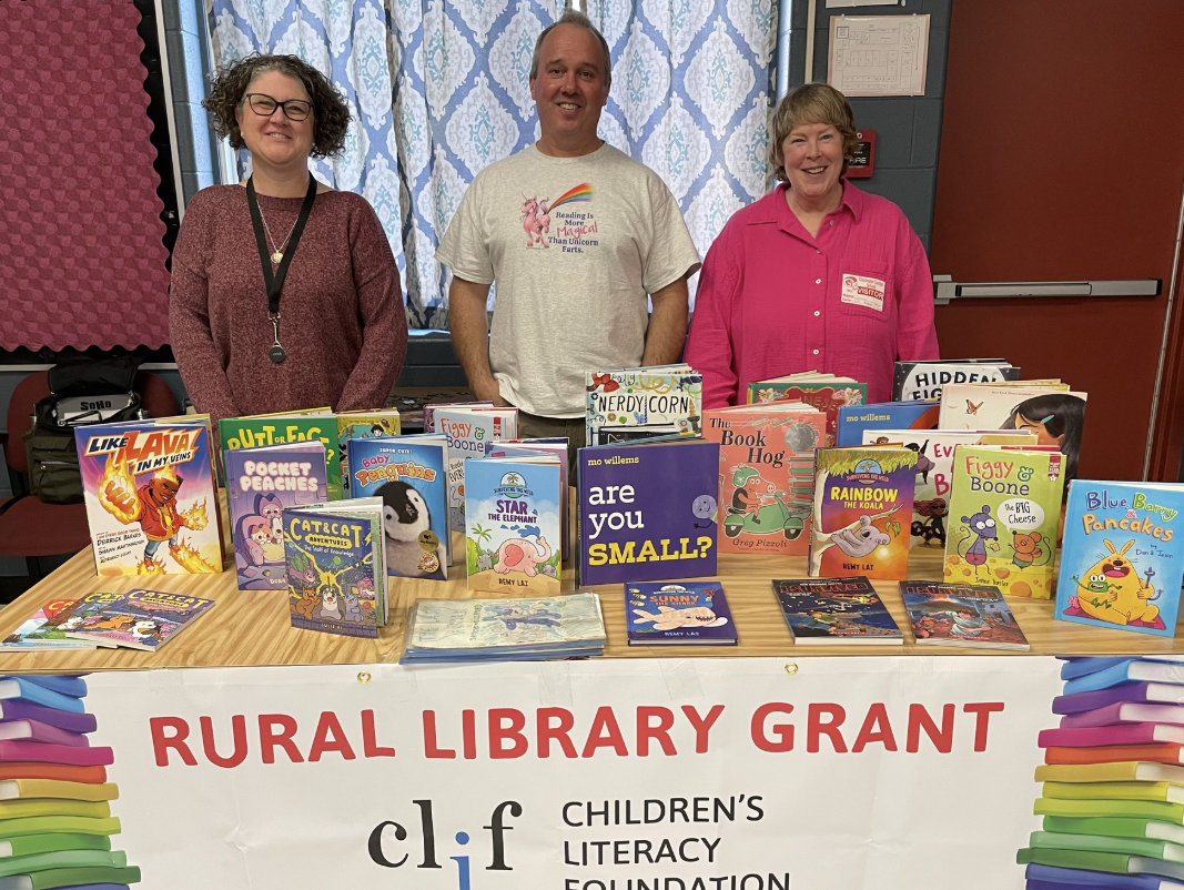 Three adults stand behind a table filled with children's books and a literacy grant banner.