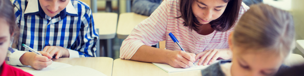 Elementary students writing in notebooks at classroom desks during a focused learning activity.