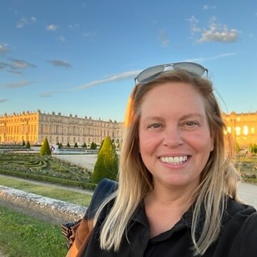 Woman smiling in front of a grand palace and manicured gardens at sunset.