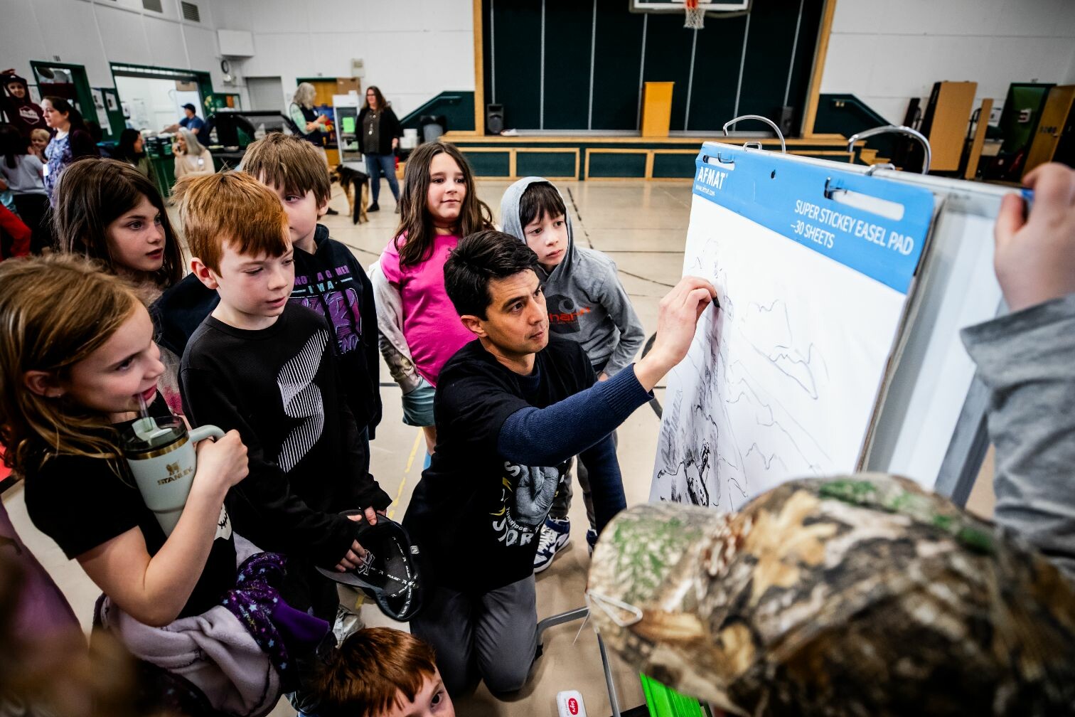 A teacher kneels by a drawing pad, explaining to captivated students in a gymnasium.