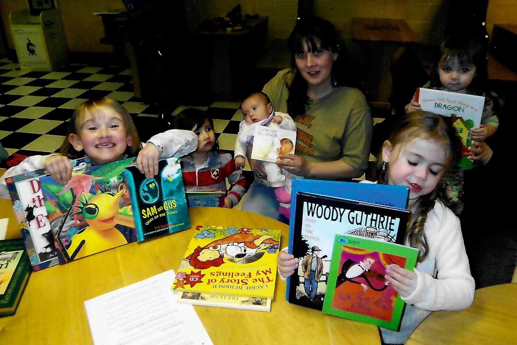 Children and a caregiver hold various children's books at a table, smiling happily.