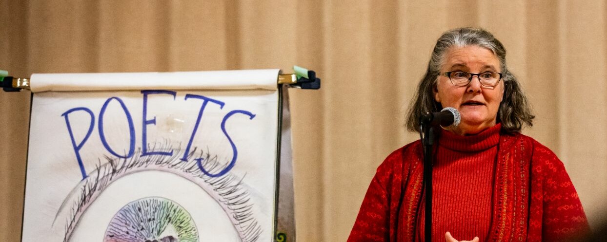 A woman in a red sweater speaks at a microphone near a "POETS" banner.