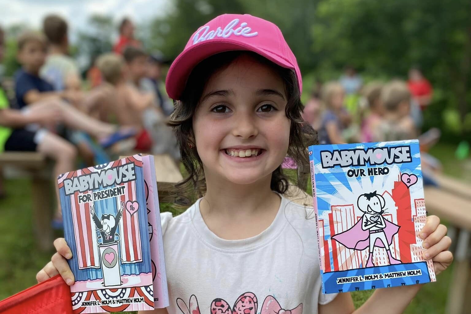 Girl in a pink cap smiles, holding two Babymouse books outdoors.
