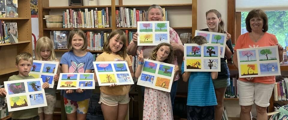 Children and adults in a library proudly display their artwork.