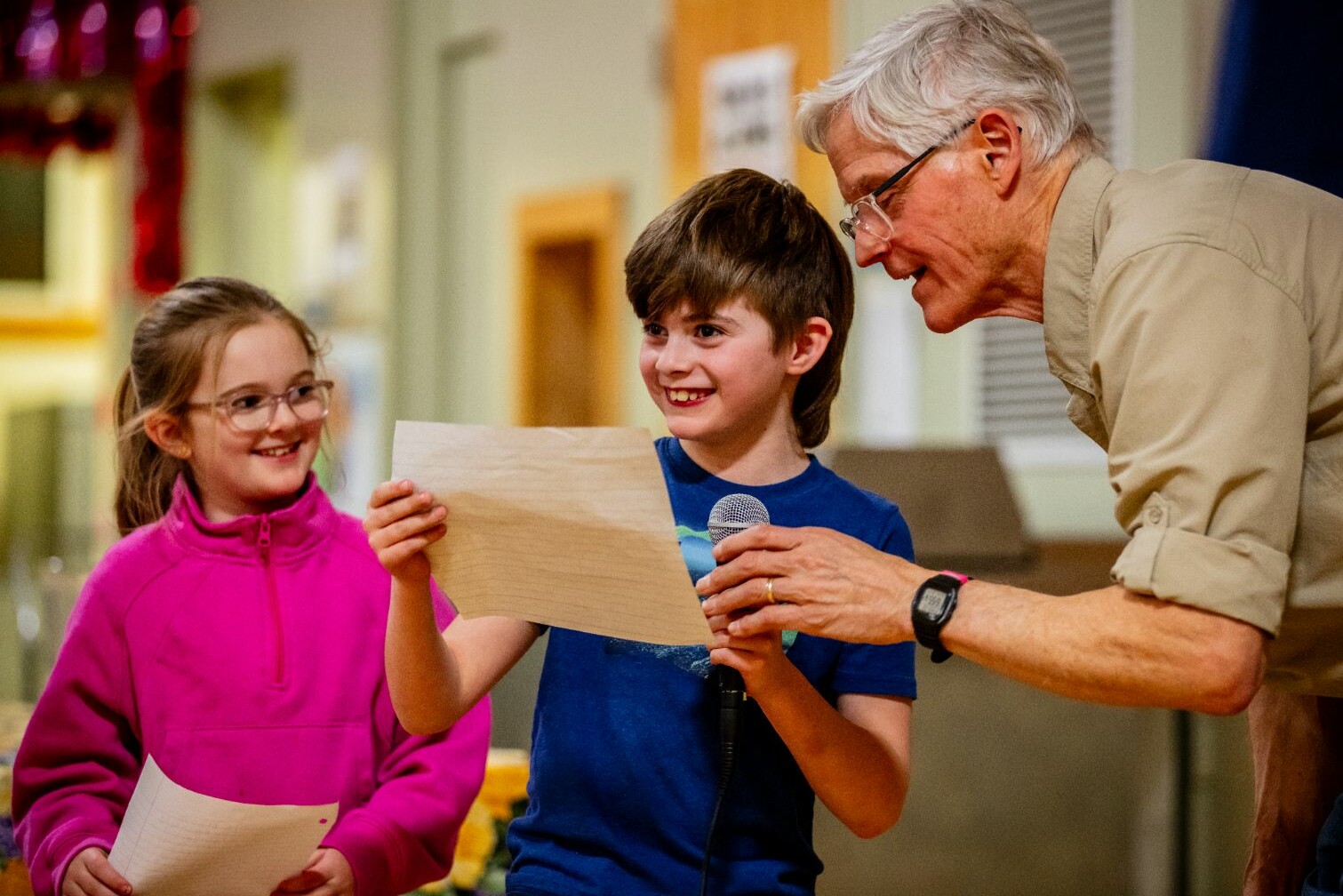 A child reads from a paper, smiling, while an adult and another child listen attentively.