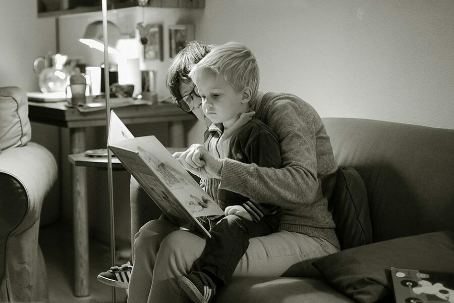 A caregiver reads a book to a young child on a couch in a cozy setting.