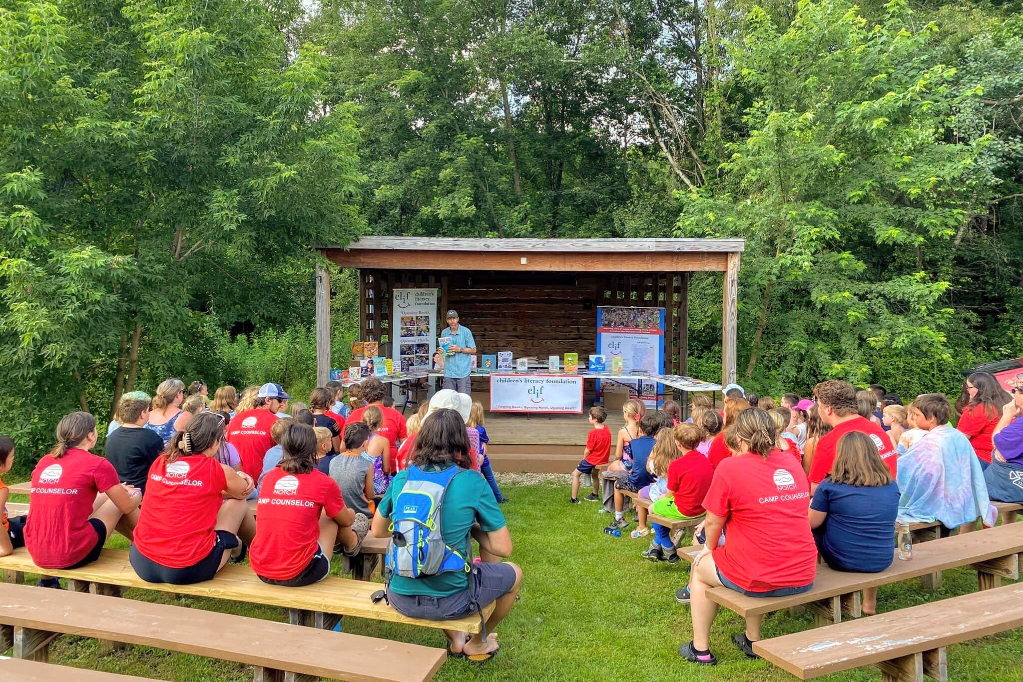A speaker addresses a group of campers seated outdoors, surrounded by trees.