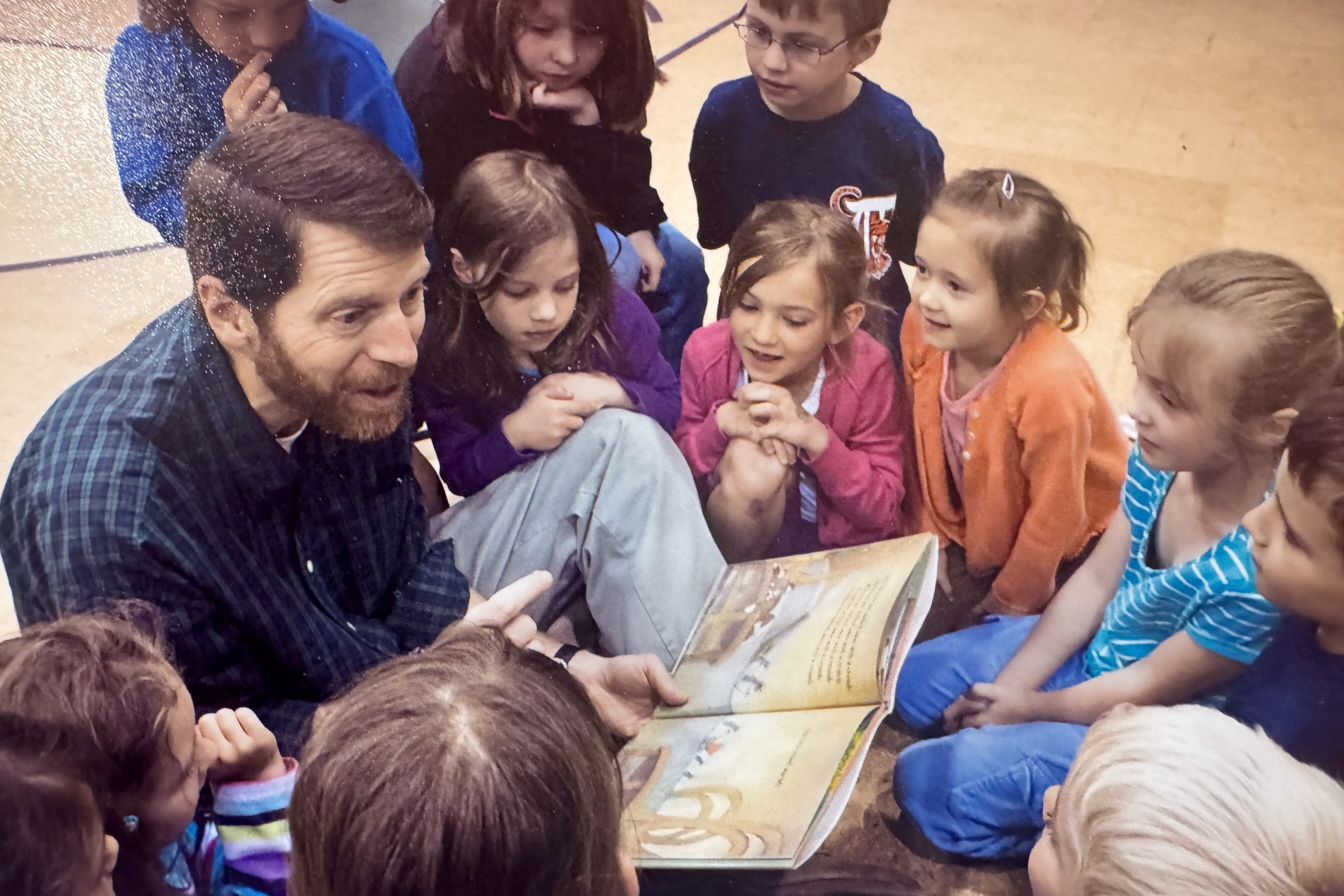 A man reads a book to a group of engaged children seated around him.