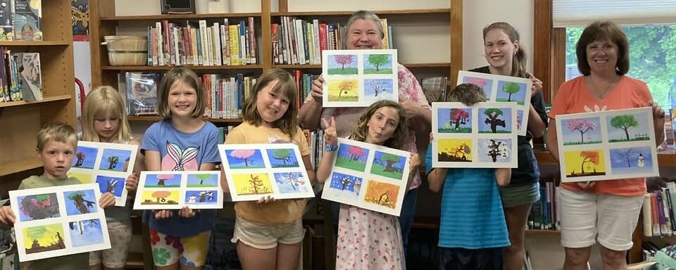 Group of children and adults holding up colorful art projects in a library setting.