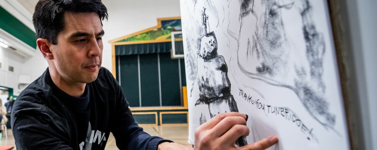 An artist sketches on a large paper while observers watch in a community space.