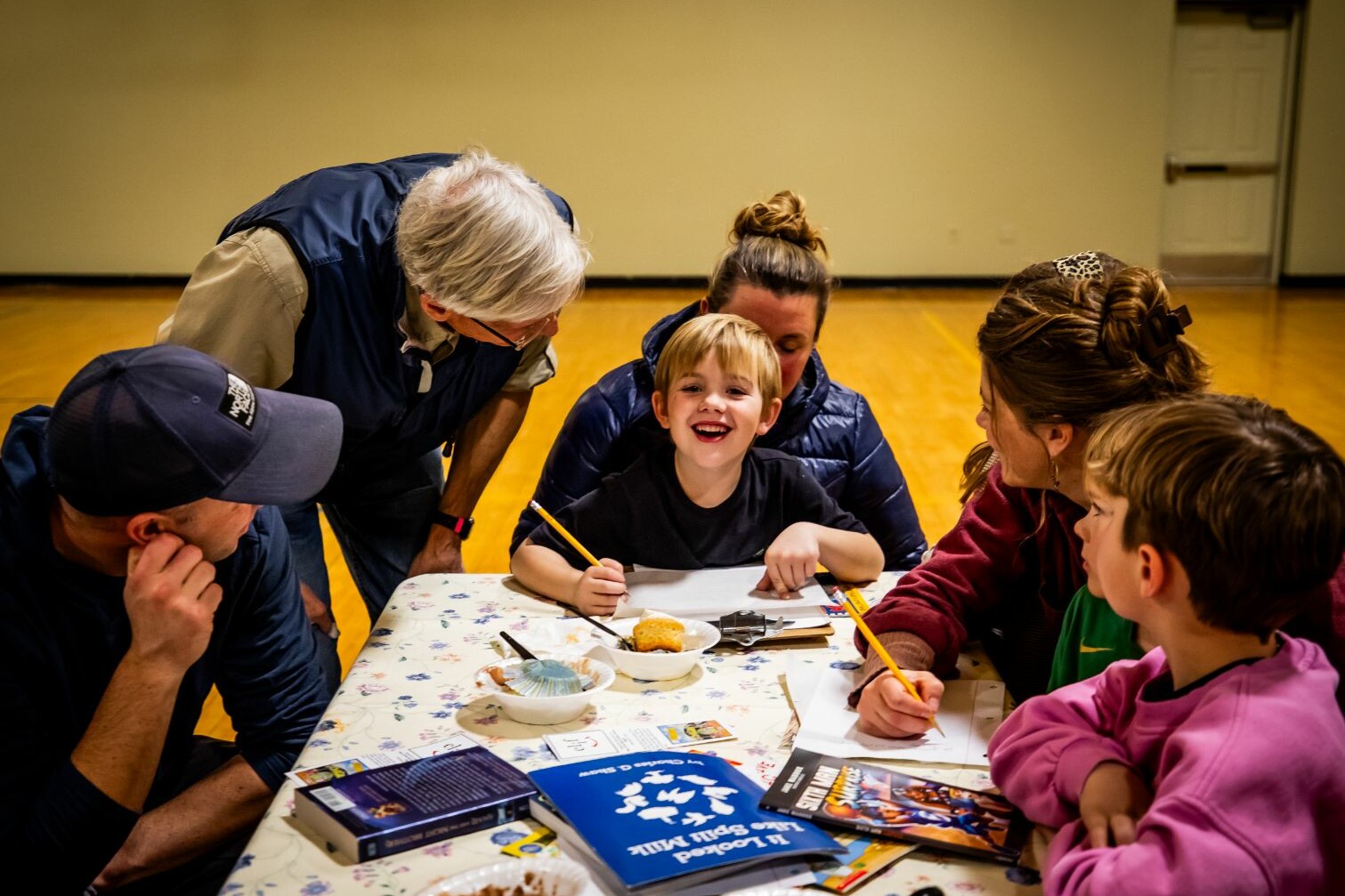 A boy smiles while drawing at a table surrounded by adults and other children.