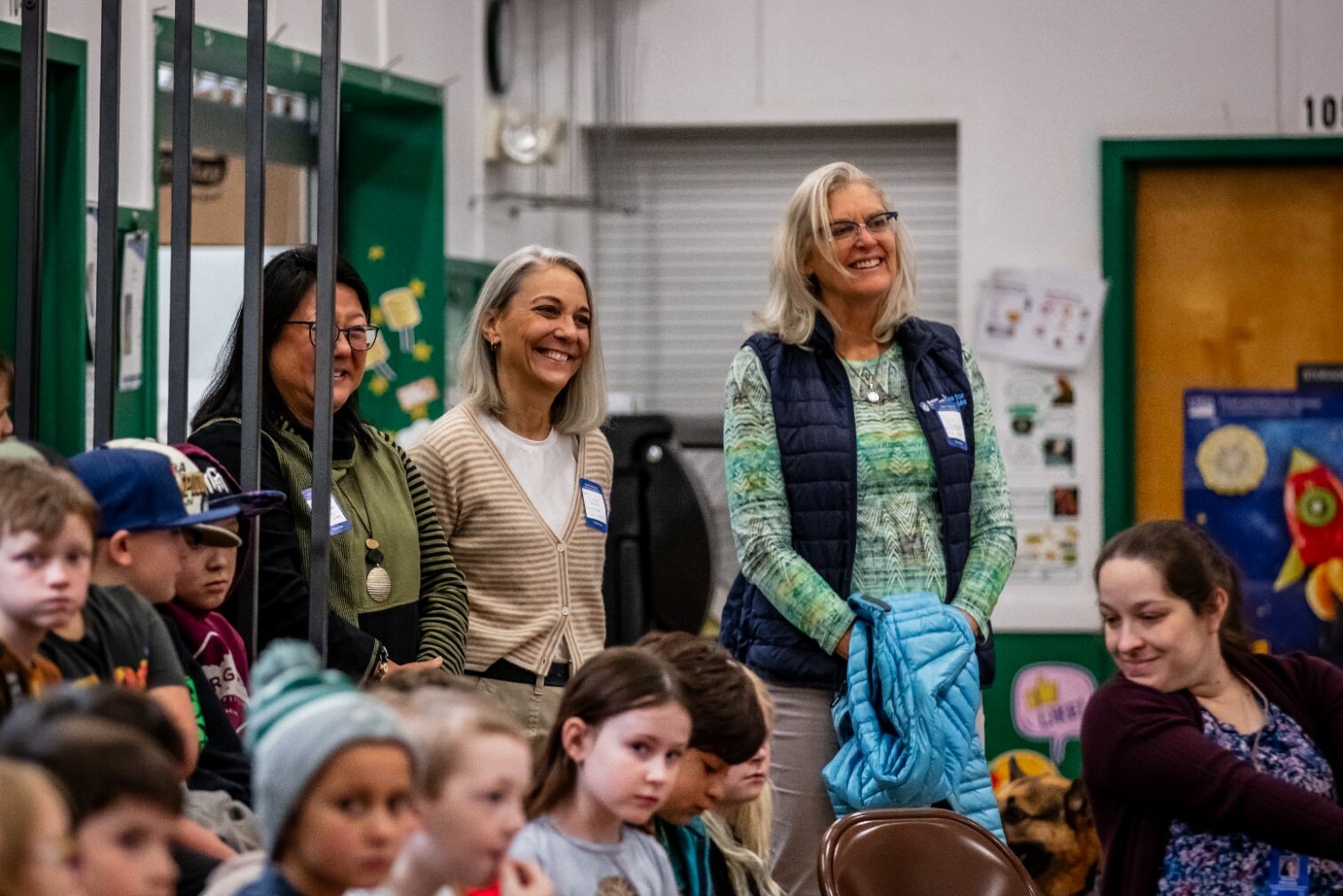 Three smiling women stand among children seated in a colorful classroom setting.