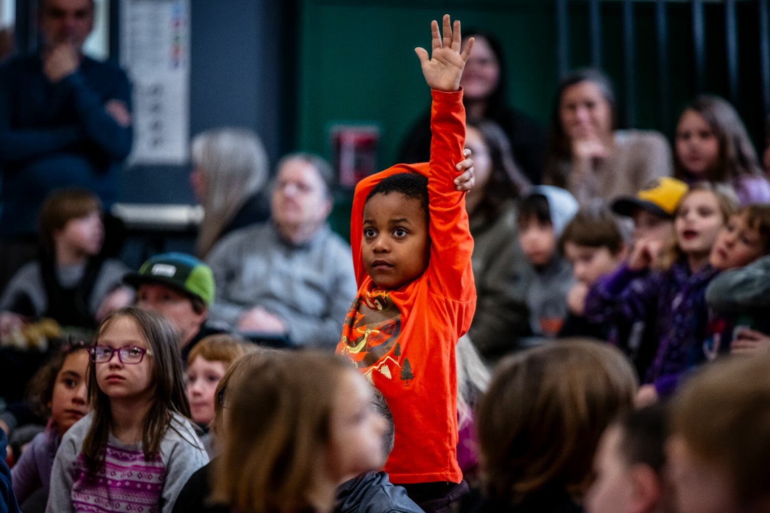 A child in an orange shirt raises their hand in a crowd of seated peers.