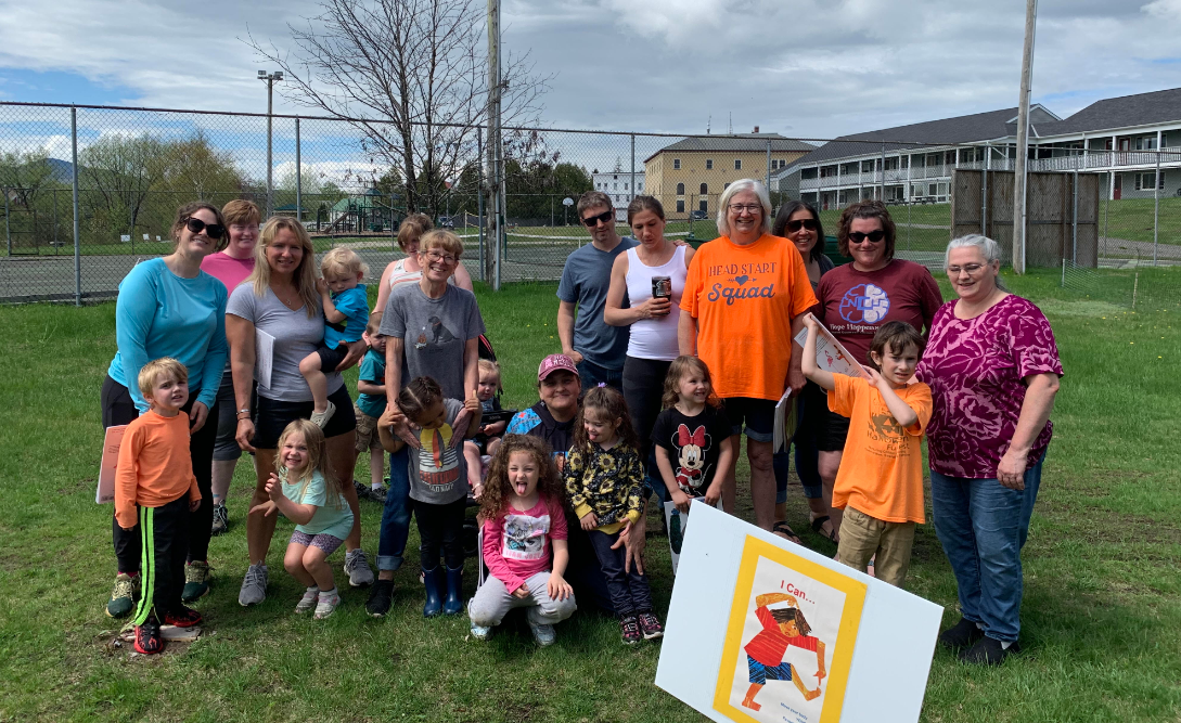Group of adults and children posing together outdoors, with a colorful sign in front.