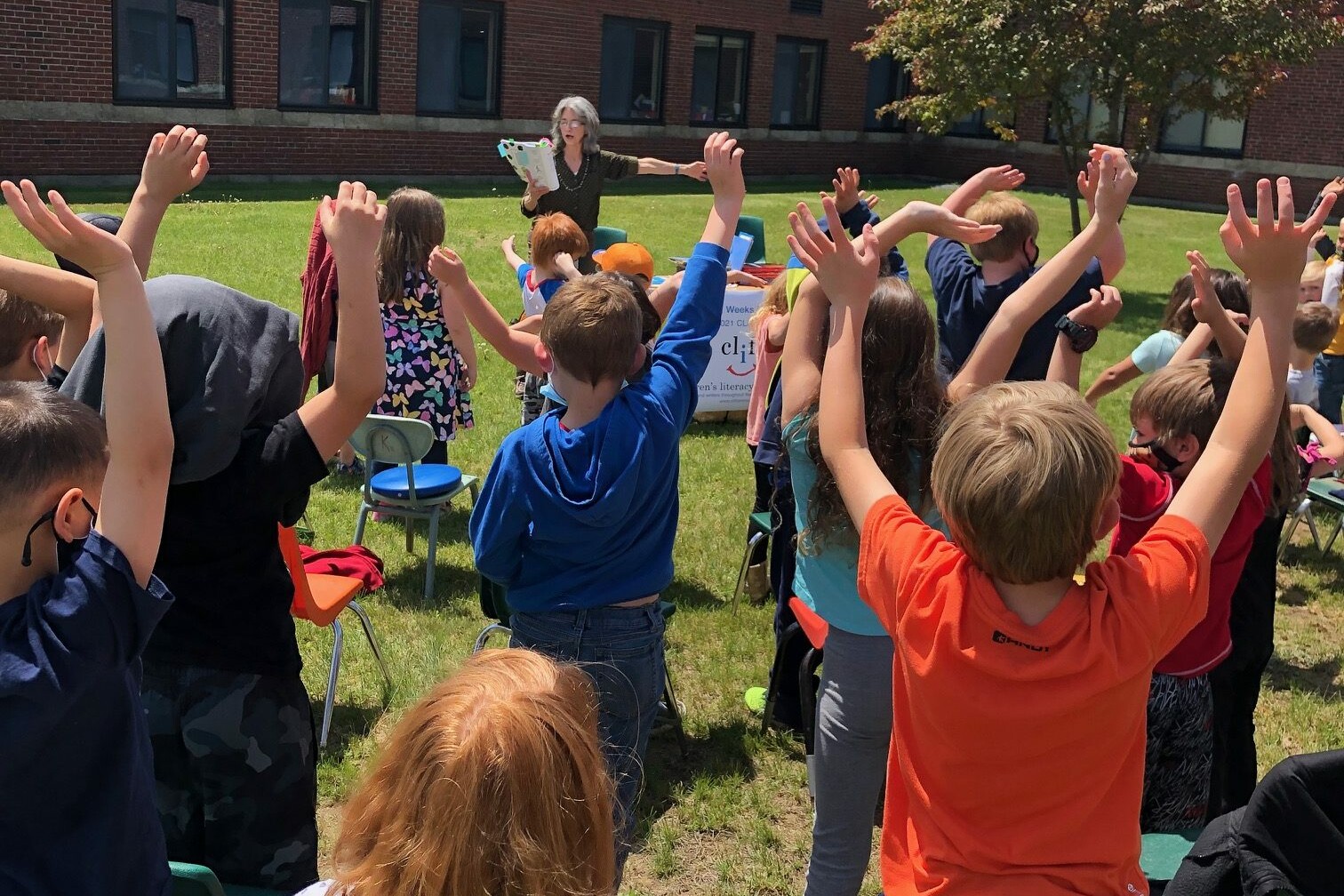 Children with raised hands listen to a presenter outdoors on a sunny day.