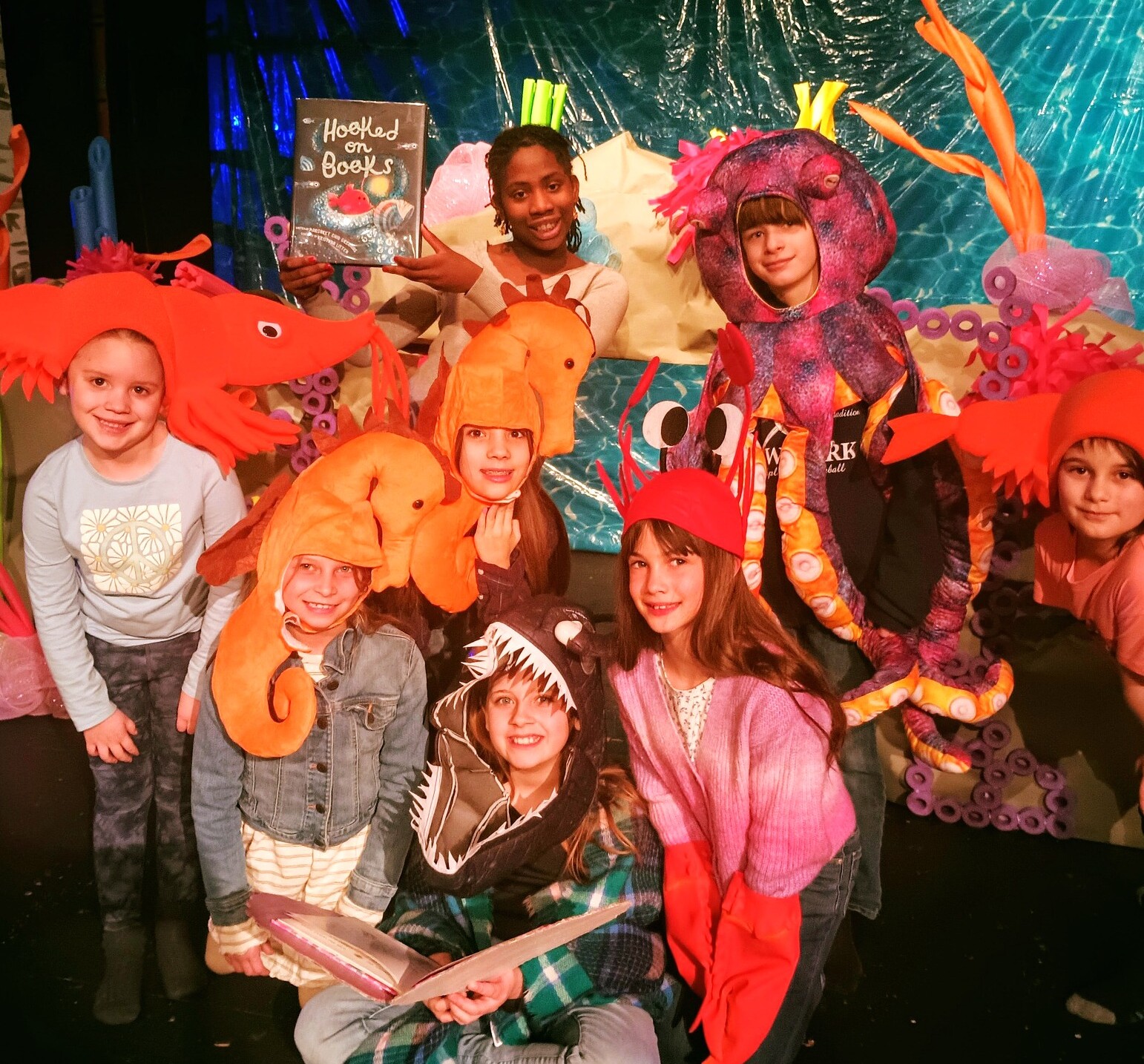 Children in colorful sea creature hats, posing with a book titled "Hooked on Books."