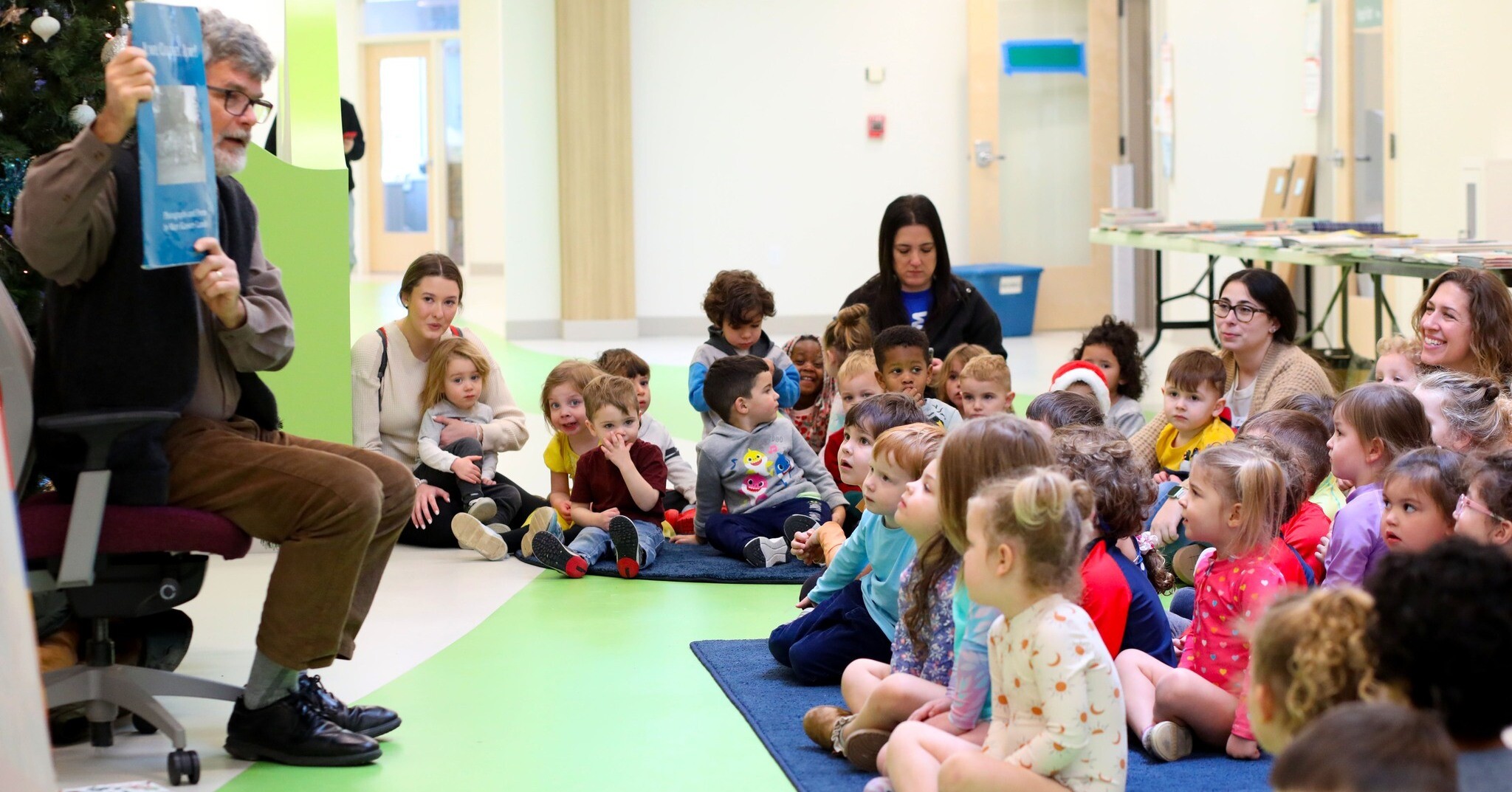 A storyteller reads to a group of engaged children and adults in a colorful space.