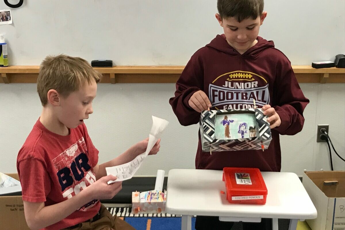 Two boys present a project, one reading a script while the other holds a decorated box.