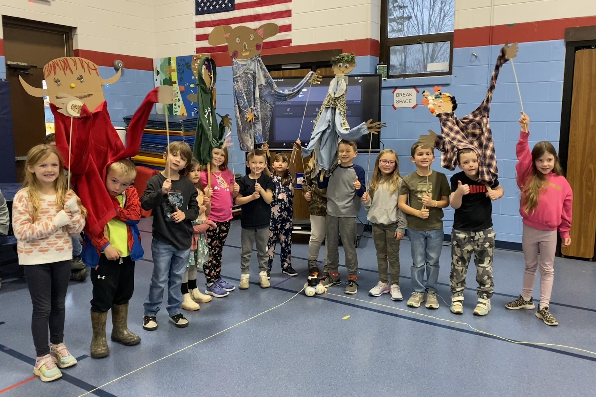 Children holding handmade puppets in a school gym, smiling and posing together.