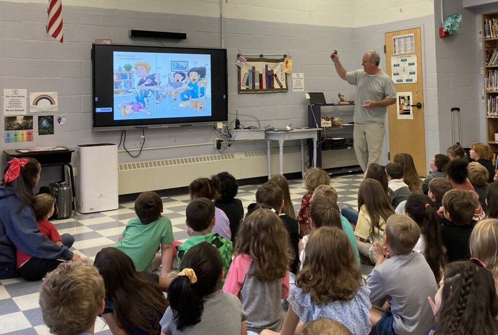 School author visit with students watching a children&rsquo;s book illustration on a screen during an interactive literacy presentation.